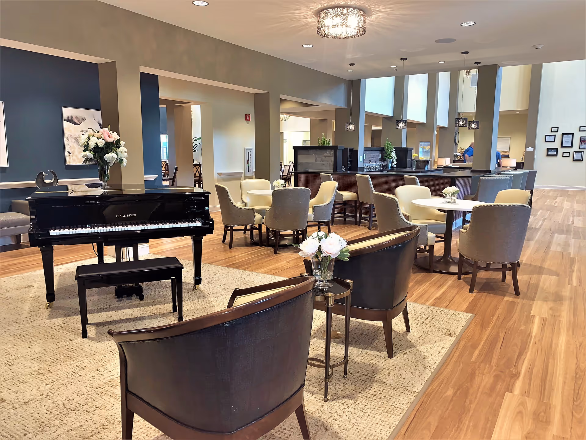 A spacious and well-lit common area in a senior living facility featuring a black Pearl River grand piano with a matching bench on a beige rug. The room has multiple seating arrangements including round tables with beige and gray upholstered chairs, and two dark wooden chairs with cream cushions near a small side table holding a vase of flowers. The floor is wooden, and the walls are painted in neutral tones with some artwork and decorative plants. Pendant lights hang from the ceiling, and there is a counter or bar area in the background.