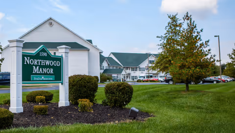 View of the exterior of Northwood Manor senior living facility with a green and white sign displaying the name and address. The building has white siding with green roofs, surrounded by a well-maintained lawn, bushes, and a tree. Several parked cars are visible in the background under a partly cloudy sky.