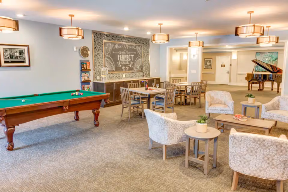 A bright communal lounge with a pool table, seating areas, dining tables, and a grand piano.