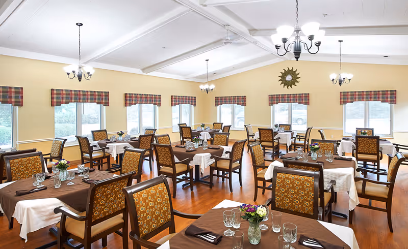 Bright dining room with multiple tables set for meals, patterned chairs, chandeliers, and windows along the walls.
