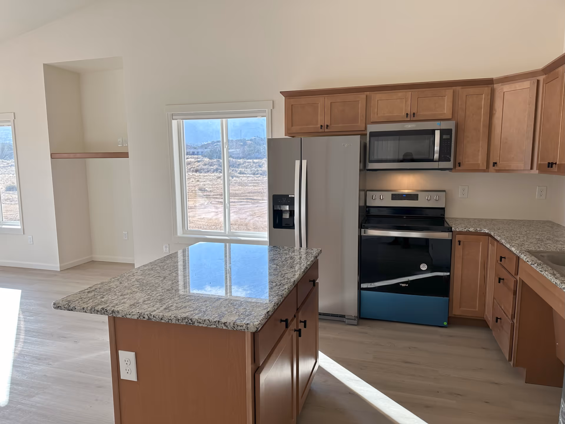 Bright kitchen with a granite island, stainless steel refrigerator, stove and microwave, and wooden cabinets with windows showing an outdoor view.