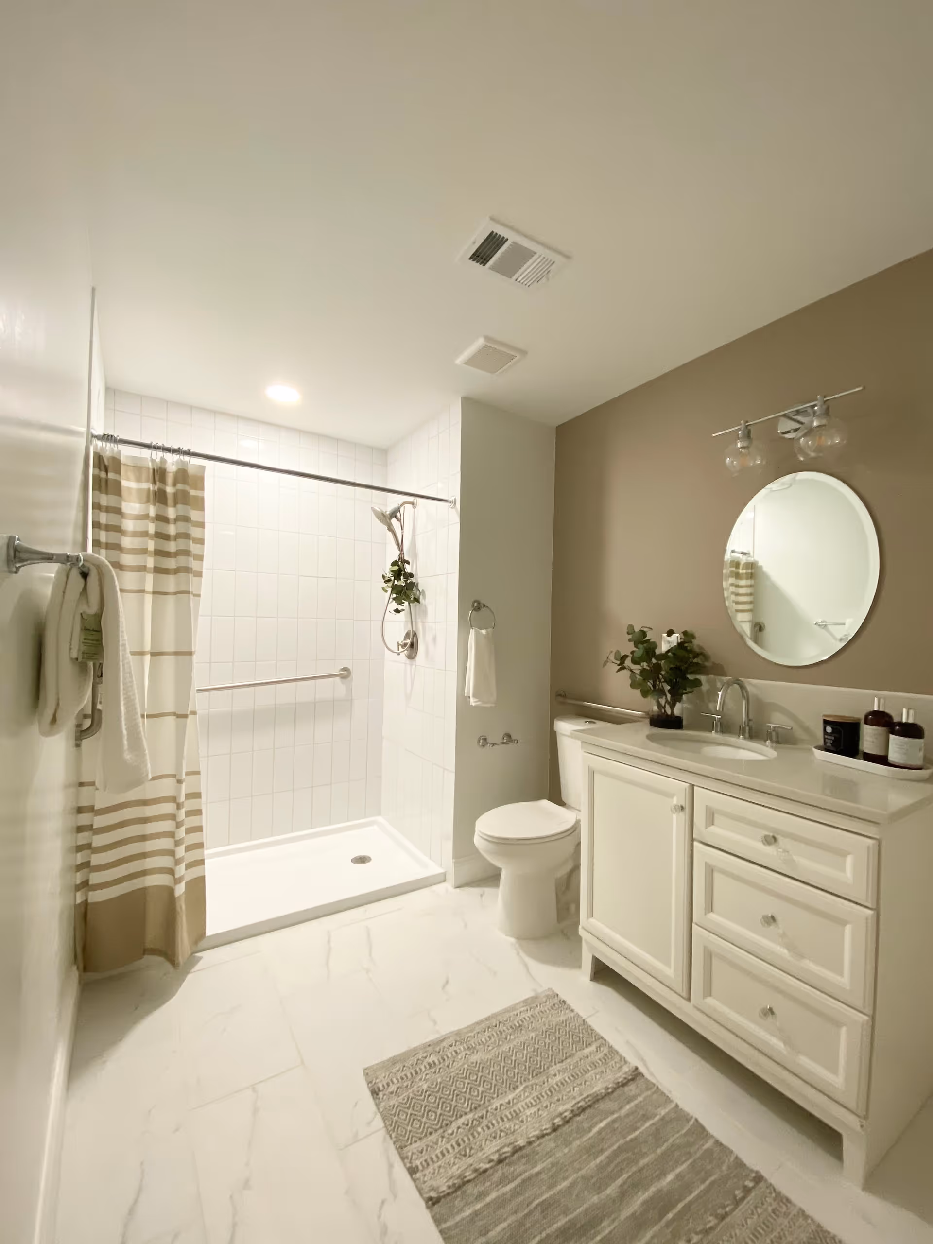 A clean and modern bathroom with a walk-in shower featuring white tiles and a beige and white striped shower curtain. There is a white toilet next to a white vanity with a sink, a round mirror above it, and two light fixtures. A small plant and toiletries are placed on the vanity. The floor is covered with white marble-like tiles and a beige patterned rug.