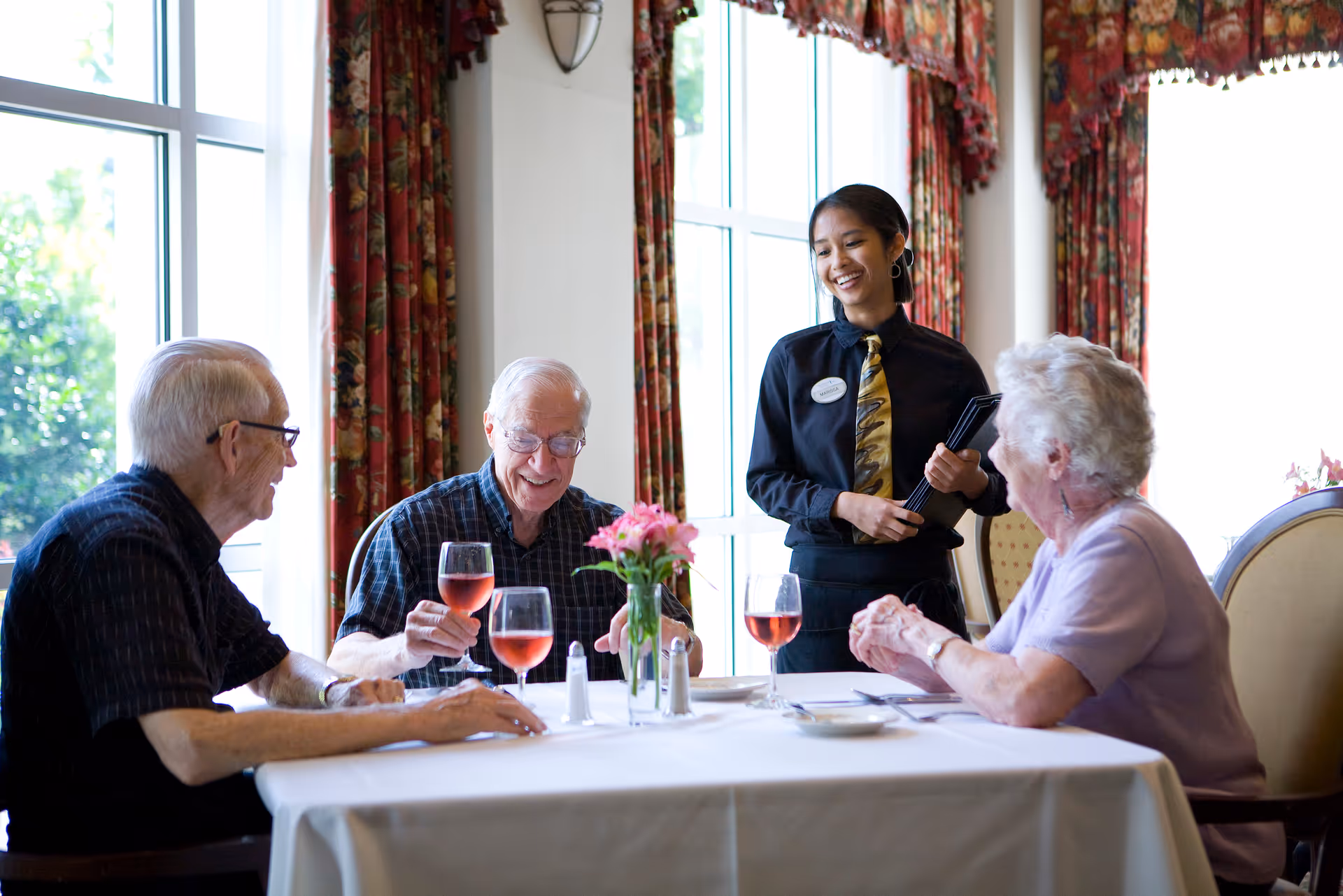 Three elderly people sitting at a dining table with glasses of rosé wine, smiling and interacting with a standing waitress who is holding a menu. The room has large windows with floral curtains and a vase with pink flowers on the table.