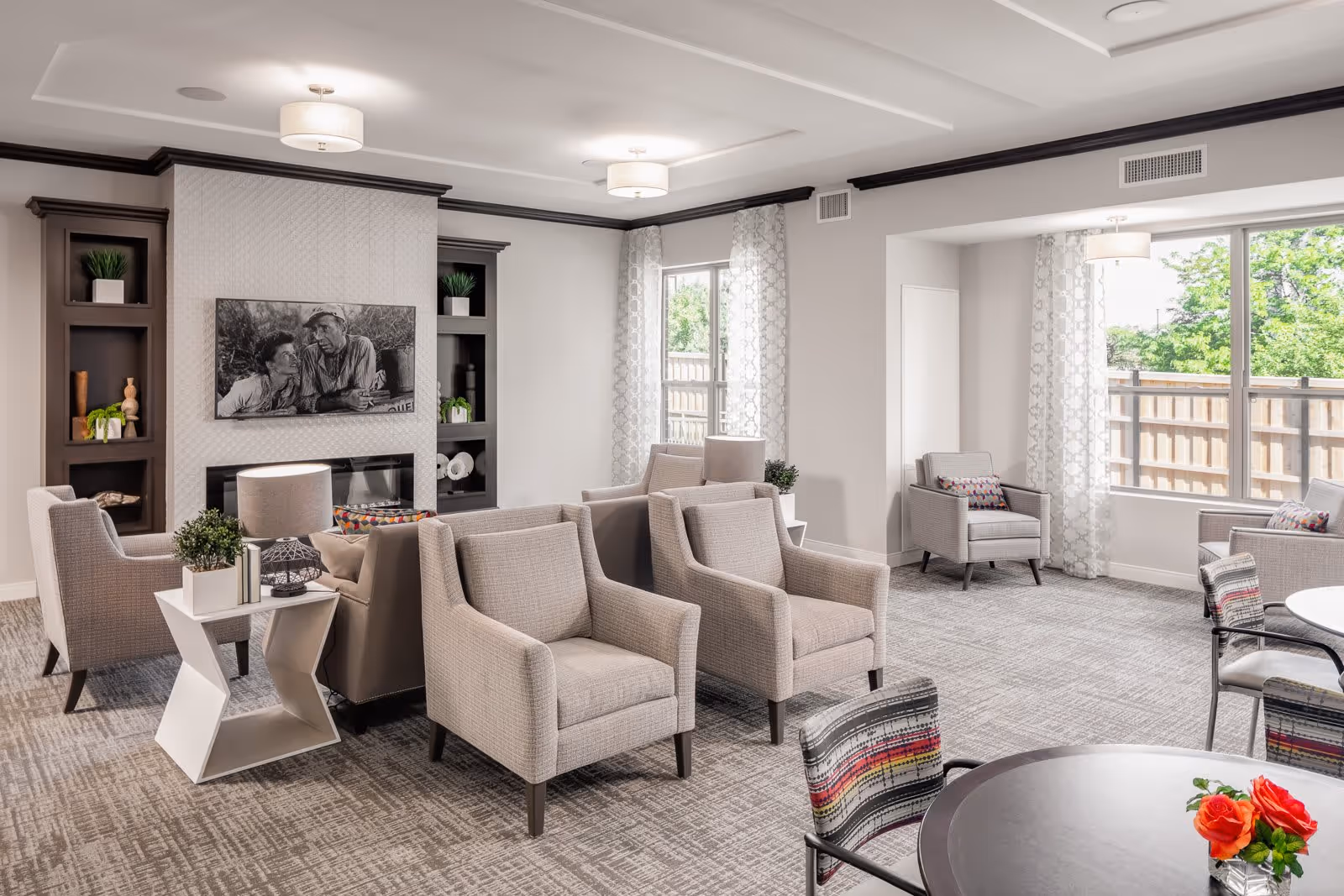 A bright and spacious living room area in a senior living facility with multiple beige armchairs arranged around a central fireplace. The fireplace is flanked by built-in shelves with decorative plants and items. Large windows with patterned curtains allow natural light to fill the room. There are also round tables with chairs, one of which has a small vase with red flowers.