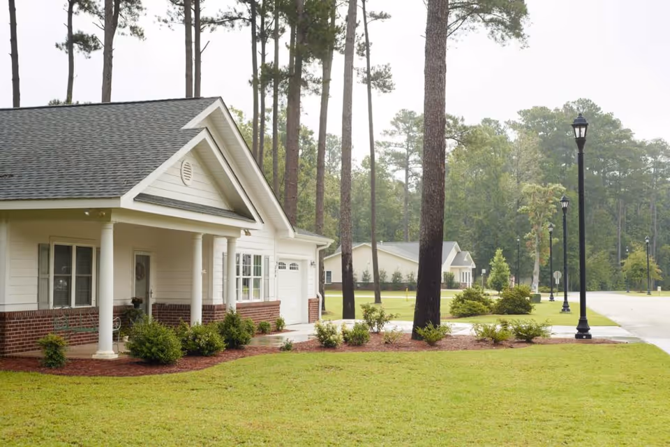 Single-story white cottage-style house with a porch and garage set among tall pine trees and manicured lawns.