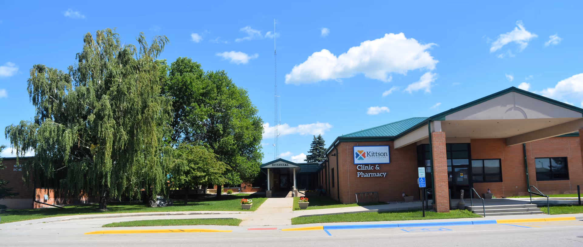 Exterior view of Kittson Healthcare building under a blue sky with scattered clouds. The building has a brick facade with a green roof and a sign that reads 'Kittson Clinic & Pharmacy'. There are trees and a sidewalk leading to the entrance, with a handicapped parking space visible in front.