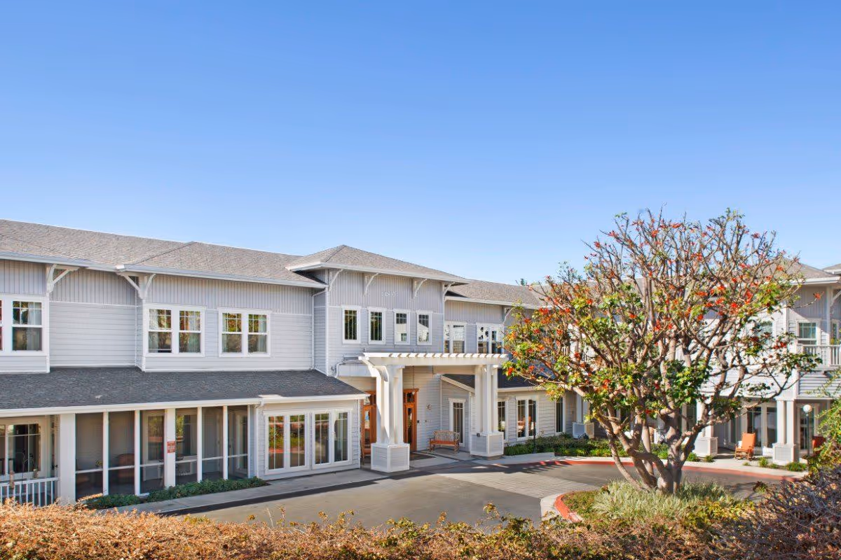 Exterior view of a two-story senior living facility building with gray siding and white trim under a clear blue sky. There is a covered entrance with white pillars and a large tree with red flowers in front of the building.