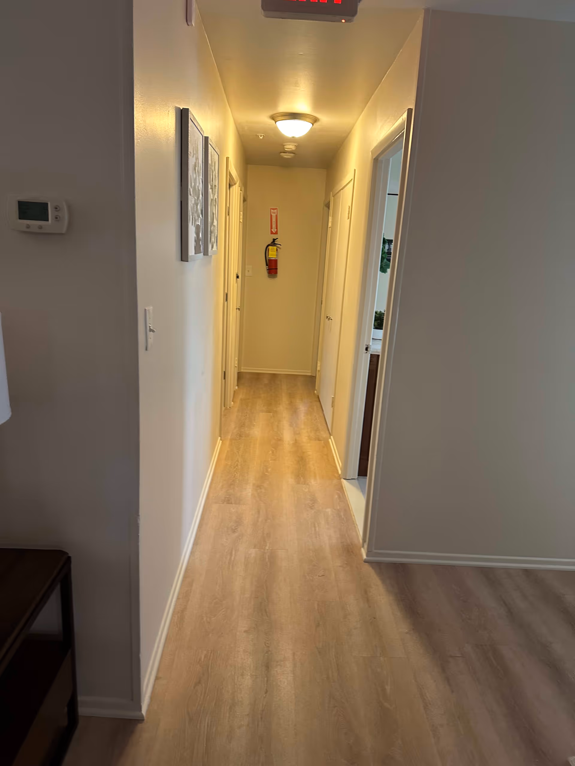 A narrow hallway with light wood flooring and cream-colored walls. There are several doors along the hallway, a fire extinguisher mounted on the far wall, and two framed pictures hanging on the left wall. A ceiling light fixture illuminates the space.