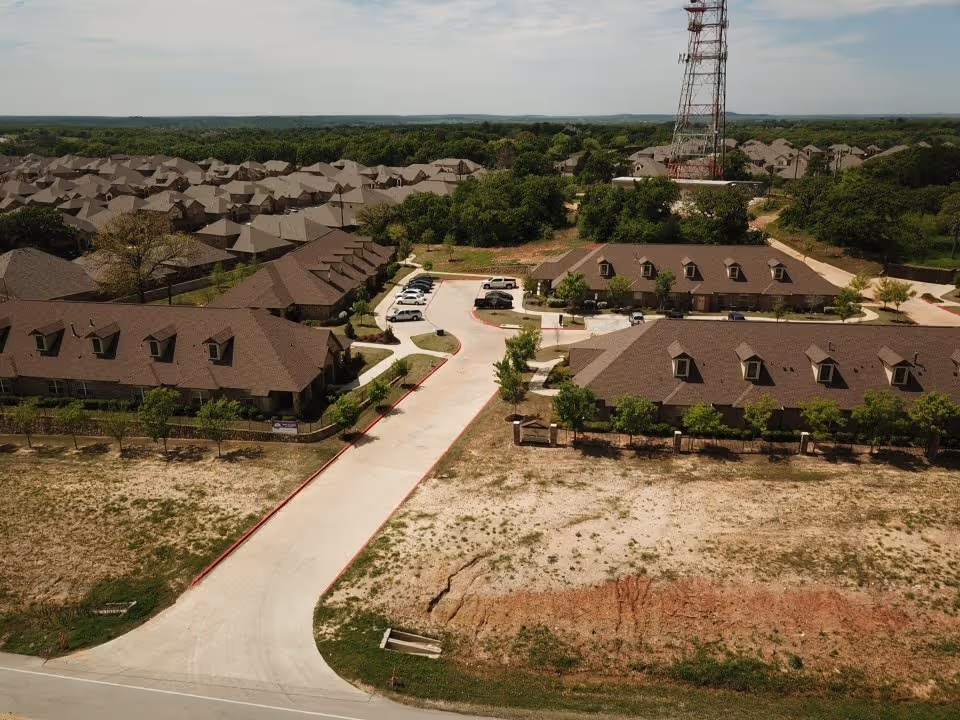 Aerial view of a residential facility with multiple brown-roofed buildings surrounded by trees and greenery. There is a paved road leading into the complex with several parked cars. A tall communication tower is visible near the center of the image.