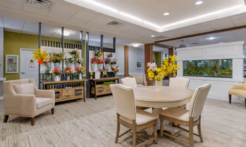 A bright and spacious common area in a senior living facility featuring a round wooden table with four upholstered chairs, two beige armchairs, and a large built-in aquarium filled with plants and fish. The room has light-colored carpeting, green accent walls, and decorative flower arrangements on shelves along one wall.