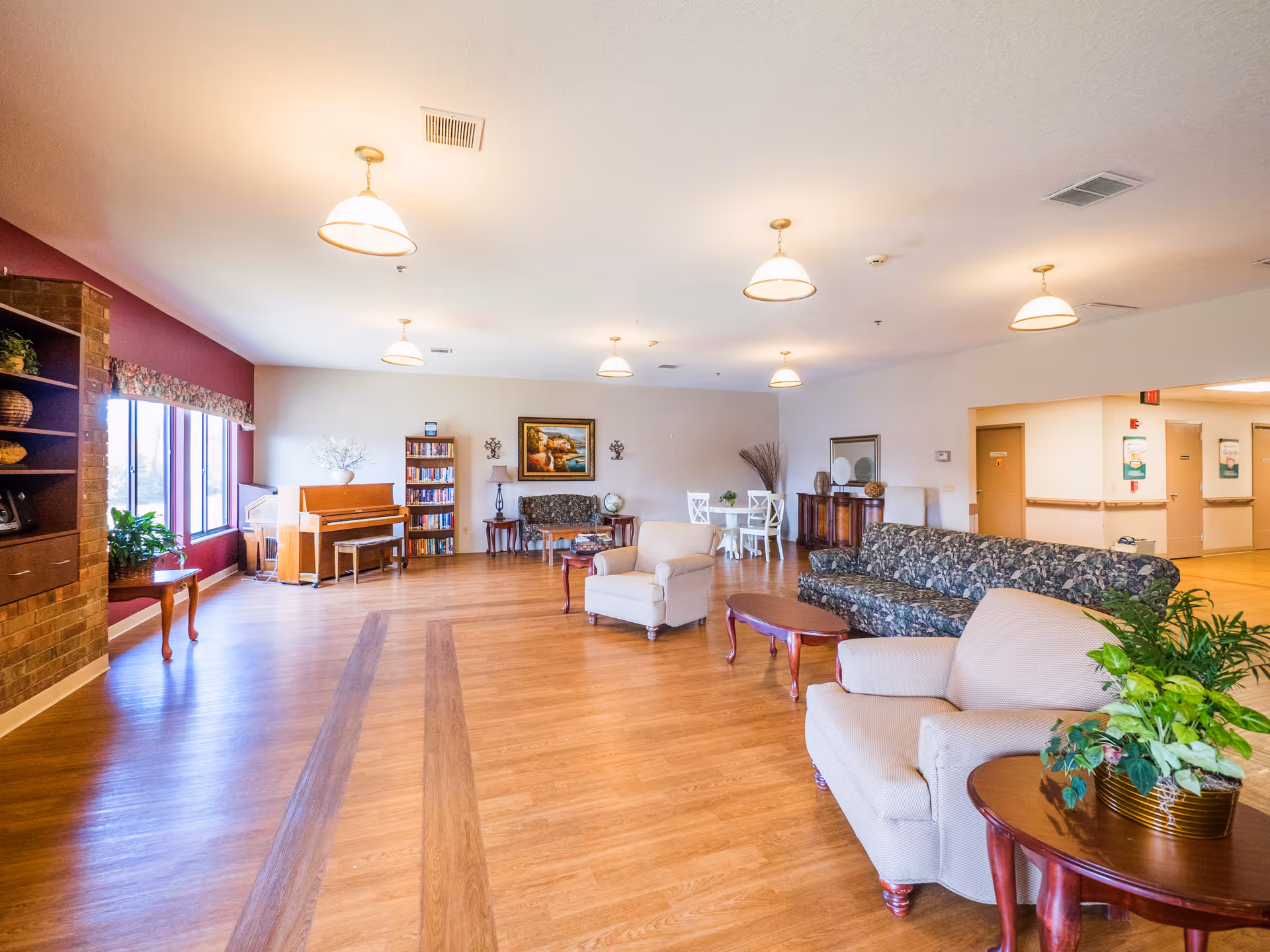 A spacious and well-lit common area in a senior living facility featuring wooden flooring, multiple armchairs and sofas with floral and plain upholstery, small wooden tables, a piano near large windows, a bookshelf, and decorative plants. The walls are painted in light colors with some artwork and wall sconces, and ceiling lights provide additional illumination.