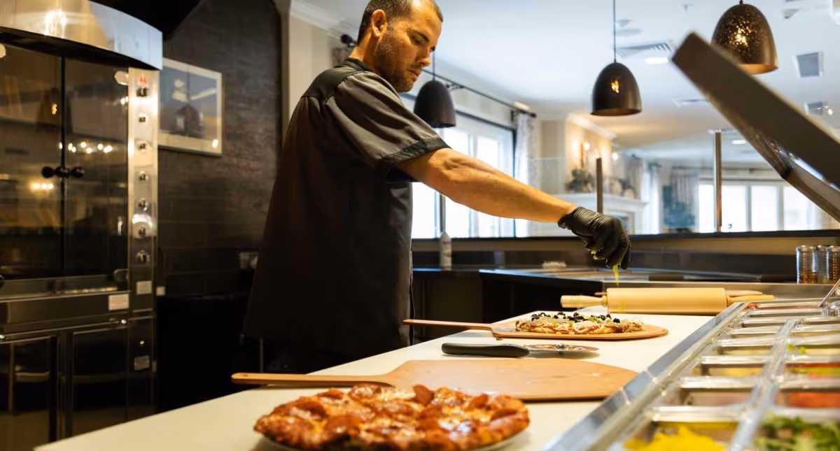 A cook in a commercial kitchen wearing gloves adds toppings to a pizza at a prep counter with ingredient bins.