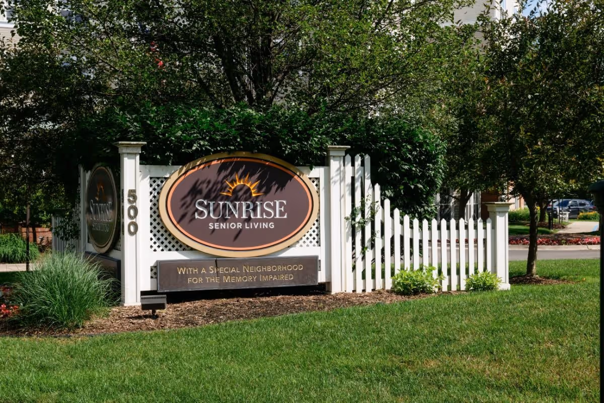 Outdoor entrance sign reading 'Sunrise Senior Living' mounted on a white picket fence with surrounding landscaping.
