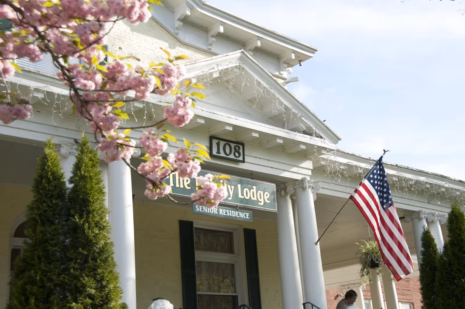 Front exterior view of a senior residence building named Ivy Lodge, featuring white columns, a porch with hanging plants, an American flag, and pink flowering tree branches in the foreground.