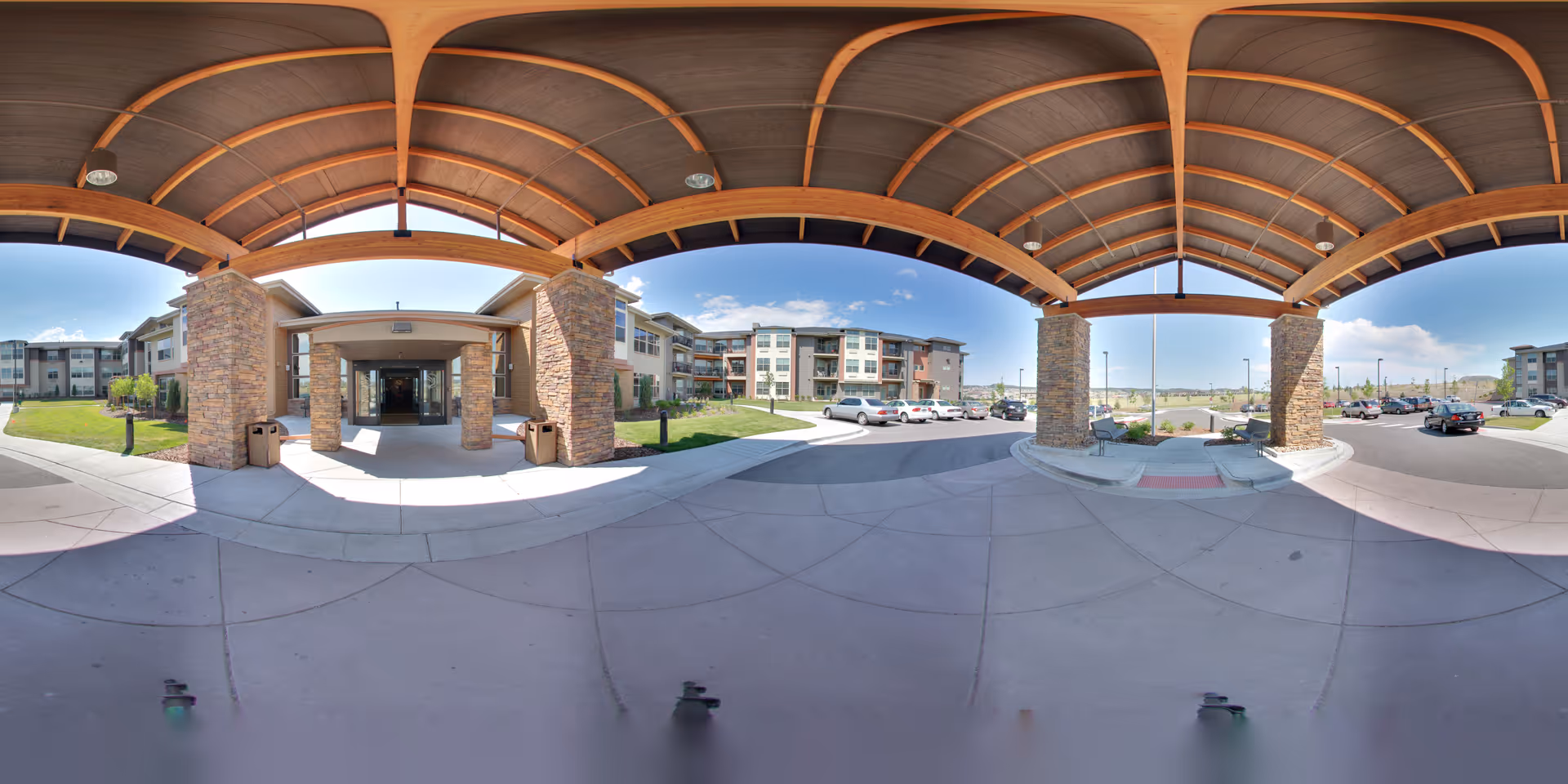 Wide-angle view of the entrance to a senior living facility with a large covered driveway supported by stone pillars and wooden beams. The building is modern with multiple floors and balconies. Several cars are parked in the parking lot under a clear blue sky.