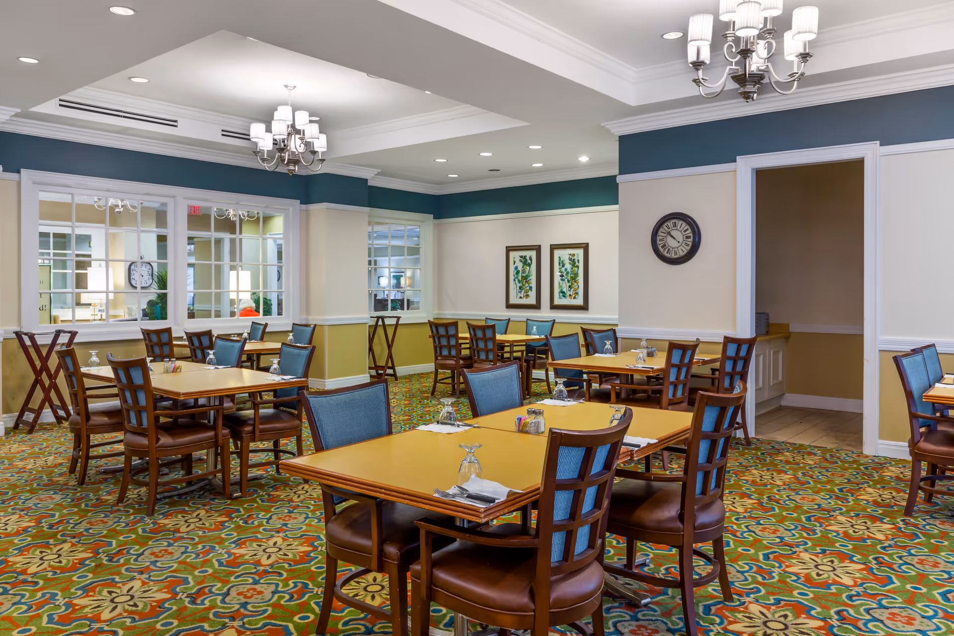 A dining room in a senior living facility with several wooden tables and chairs arranged neatly. The tables are set with upside-down glasses, napkins, and silverware. The room features a colorful patterned carpet, light-colored walls with a blue accent near the ceiling, and decorative framed artwork on the walls. Two chandeliers hang from the ceiling, and there is a clock on one wall. A doorway leads to another room.