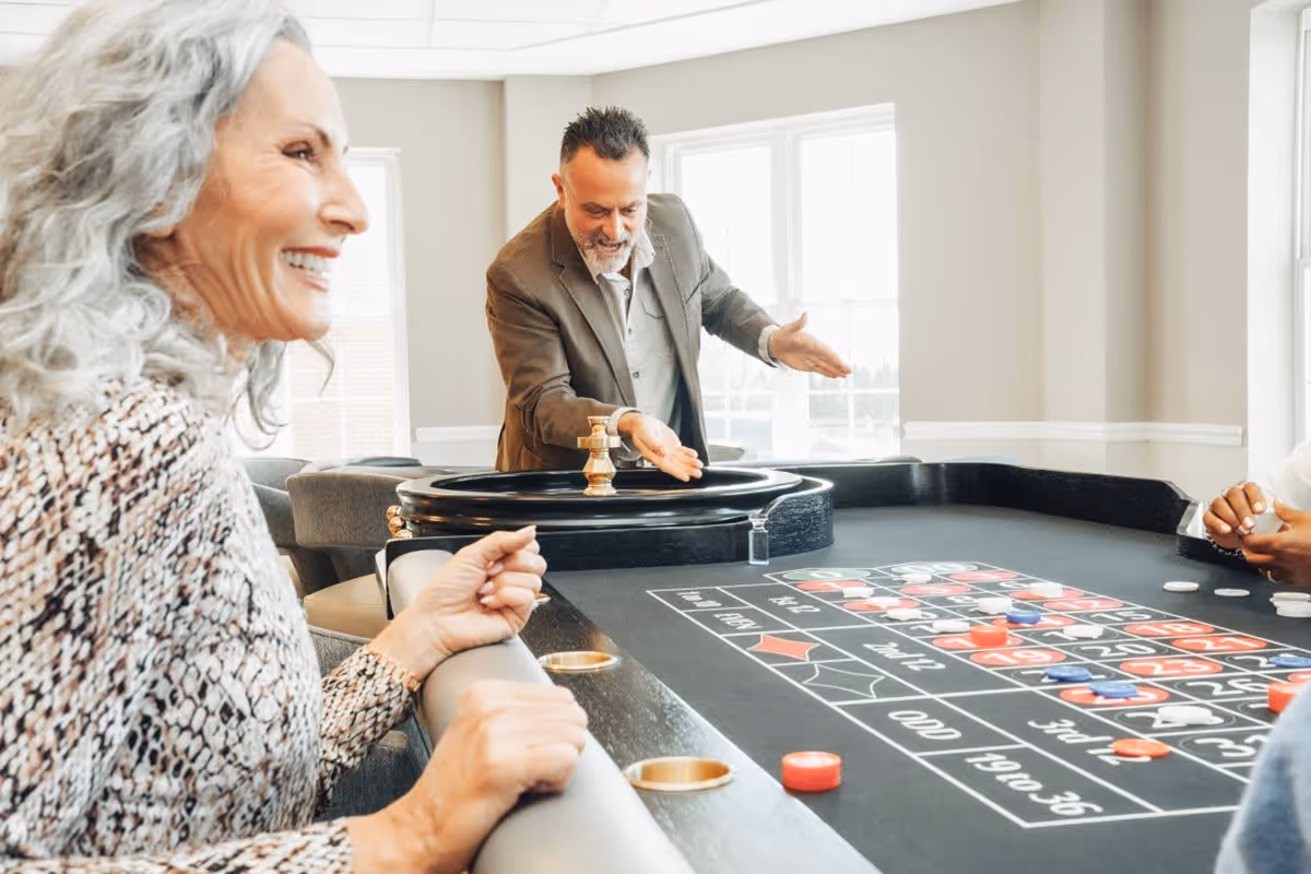 A smiling elderly woman and a man in a suit playing roulette at a casino-style gaming table in a bright room with large windows.