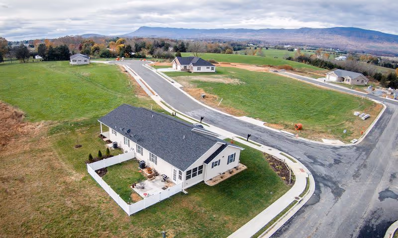 Aerial view of a single-story residential building with a fenced backyard, surrounding roads and grassy lots in a suburban development with hills in the background.