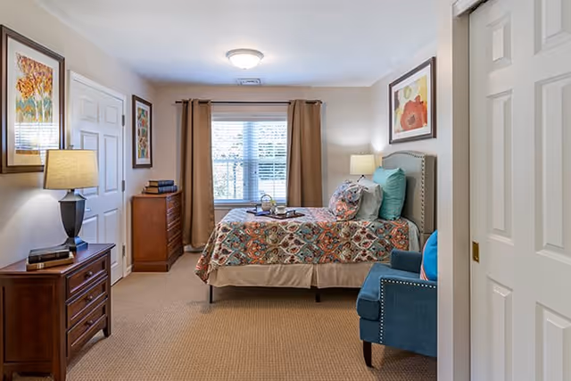 A cozy bedroom in an assisted living facility featuring a bed with a colorful patterned bedspread and multiple pillows. The room includes a wooden dresser with a lamp and books, a blue upholstered chair, framed artwork on the walls, beige curtains covering a window, and a carpeted floor.