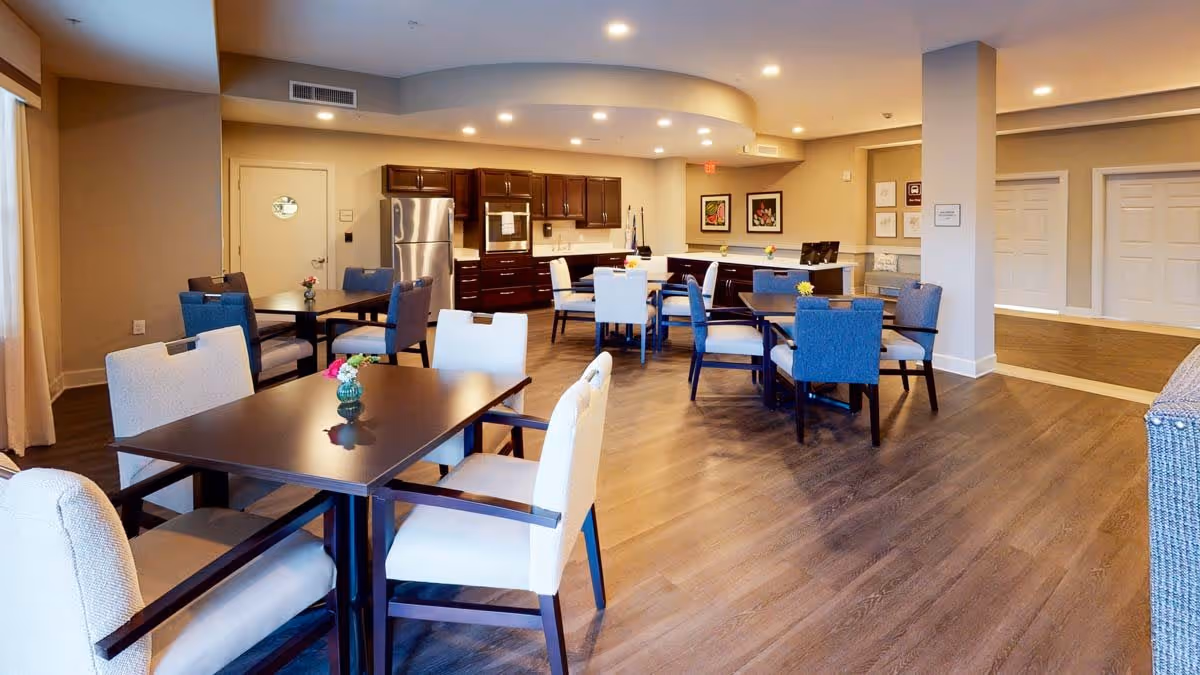 Communal dining area with multiple tables and upholstered chairs and a kitchen counter in the background.