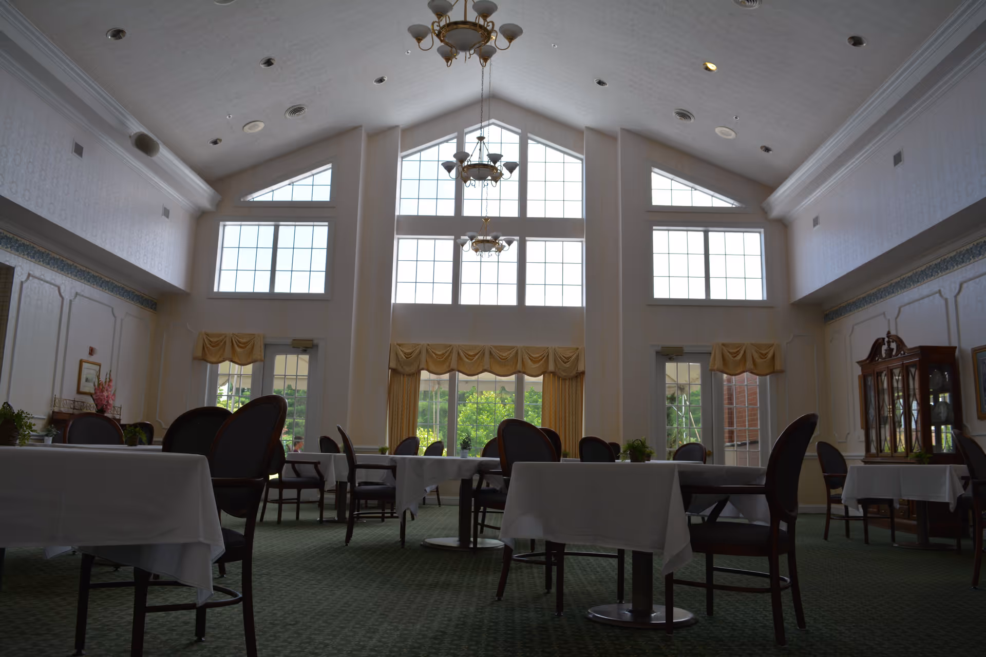 A spacious dining room with large windows allowing natural light to fill the space. The room features multiple tables covered with white tablecloths and surrounded by dark wooden chairs. The high ceiling has elegant chandeliers, and there are yellow curtains on the windows. The walls are decorated with molding and a display cabinet is visible on the right side.