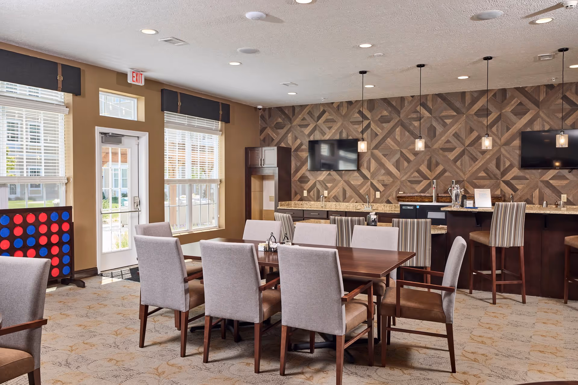 A well-lit common area with a large wooden table surrounded by cushioned chairs. The room features a patterned wooden accent wall with two mounted flat-screen TVs, a countertop with bar stools, pendant lights hanging from the ceiling, and large windows letting in natural light. A large Connect Four game is visible near the exit door.