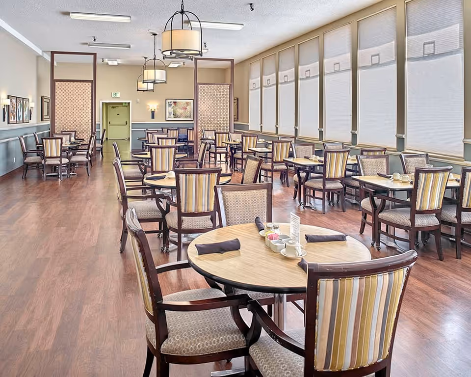 Bright dining room with round tables and striped chairs arranged on a wood floor beside large windows.