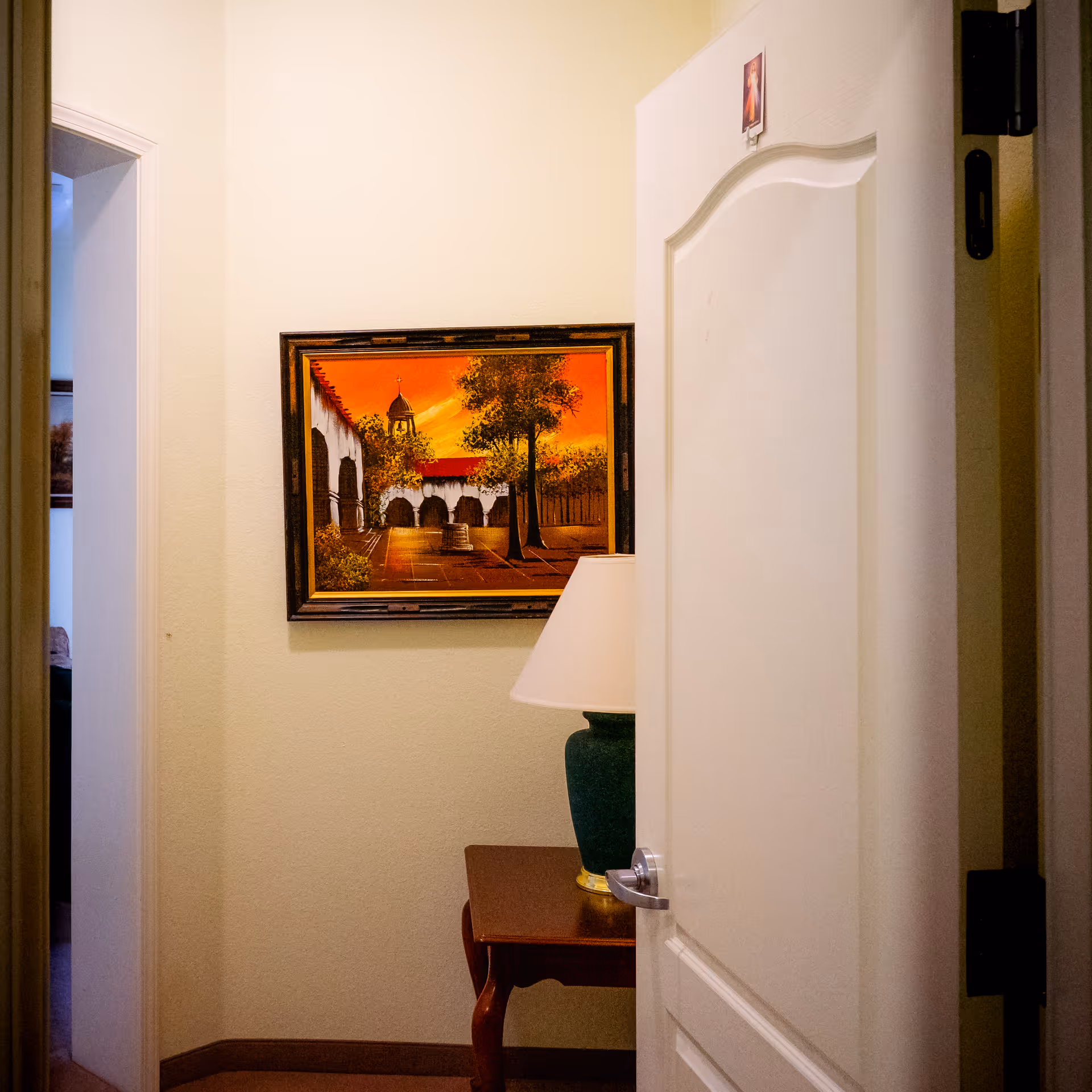Partially open white door revealing a small corner with a wooden table holding a green lamp with a white lampshade. On the wall above the table is a framed painting depicting an outdoor scene with trees and a building under an orange sky.