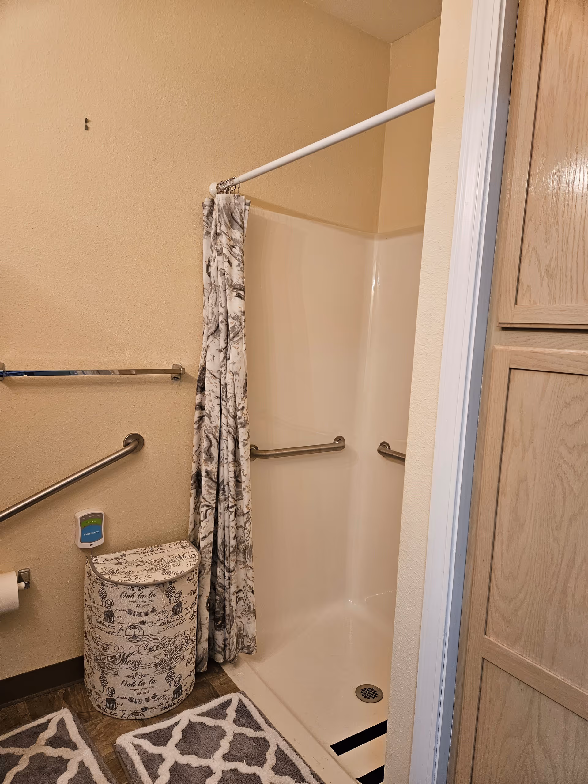 A bathroom shower area with beige walls and a white shower stall. The shower has a patterned curtain partially drawn to the side and two metal grab bars inside. Outside the shower, there is a decorative laundry hamper with script writing, a toilet paper holder with a roll of toilet paper, and two gray and white patterned rugs on the floor. A wooden cabinet is visible on the right side of the image.
