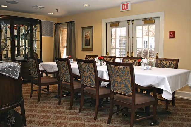 A formal dining room with a long table covered in a white tablecloth, upholstered wooden chairs, small floral centerpieces, a china cabinet, and double doors beneath an EXIT sign.