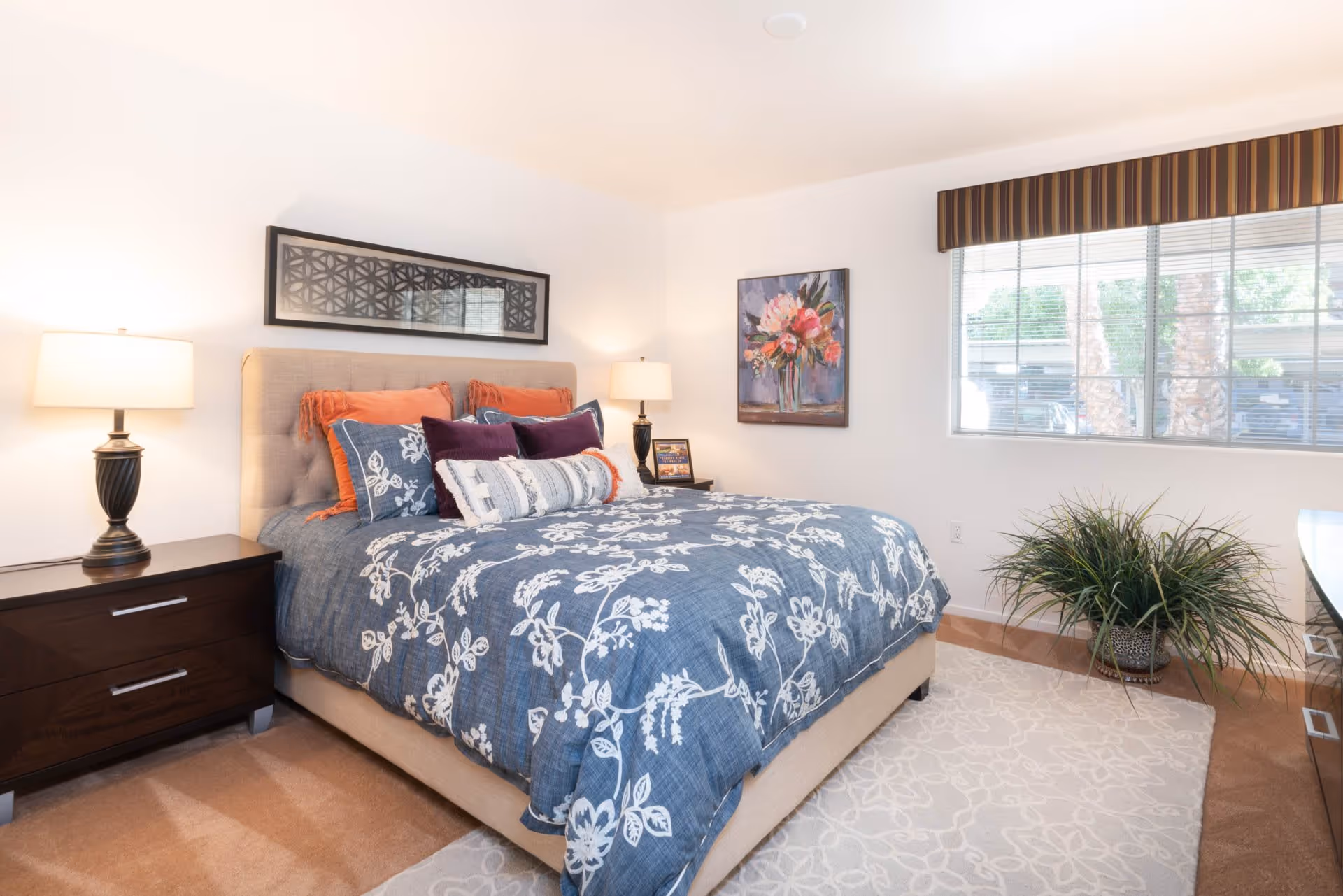 A bright bedroom with a large bed featuring a blue floral comforter and multiple pillows. There are two dark wooden nightstands on either side of the bed, each with a lamp. A framed artwork hangs above the bed, and another floral painting is on the adjacent wall. A large window with striped valance lets in natural light, and a green potted plant sits near the window on a light-colored rug.