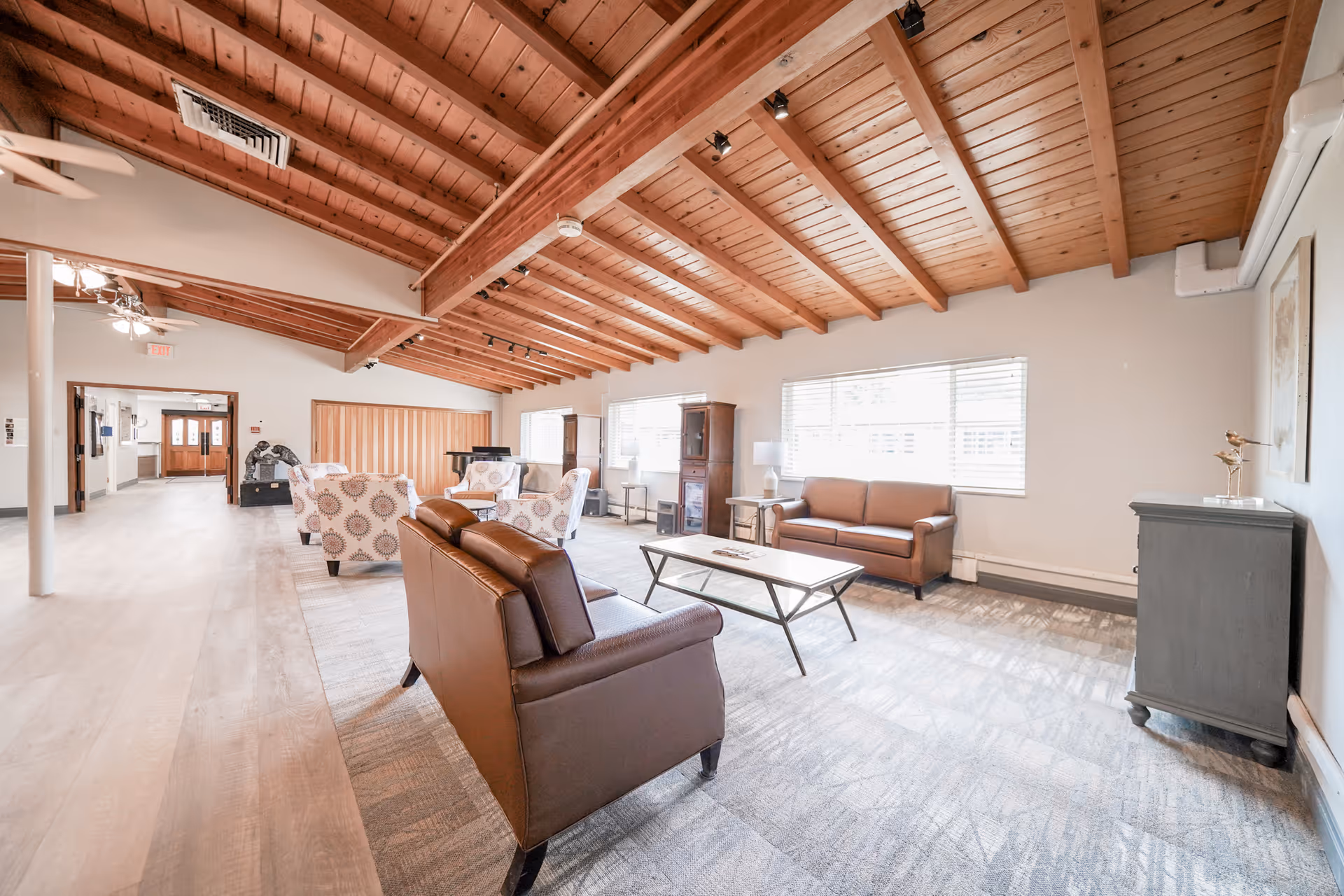A spacious living room area in a nursing and rehabilitation center with a high wooden beam ceiling and large windows letting in natural light. The room features brown leather sofas, patterned armchairs, a coffee table, a cabinet, and a side table with a lamp. The floor is covered with a light-colored carpet and wood flooring, and the space looks clean and inviting.