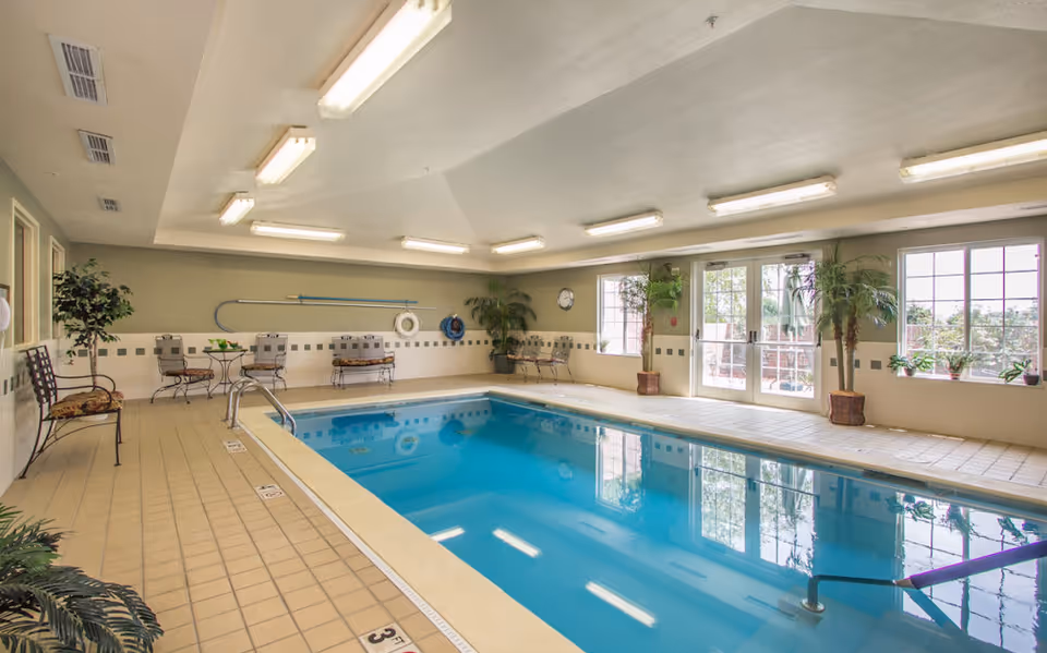 Indoor swimming pool area with tiled floor, several metal chairs with cushions, potted plants, large windows and glass doors letting in natural light, and ceiling lights.