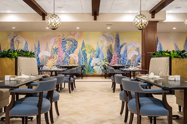 Dining room with multiple tables and blue upholstered chairs arranged neatly. The walls feature a colorful mural of abstract floral designs. Pendant lights hang from the ceiling with wooden beams, and there are green plants on top of partition walls between seating areas.