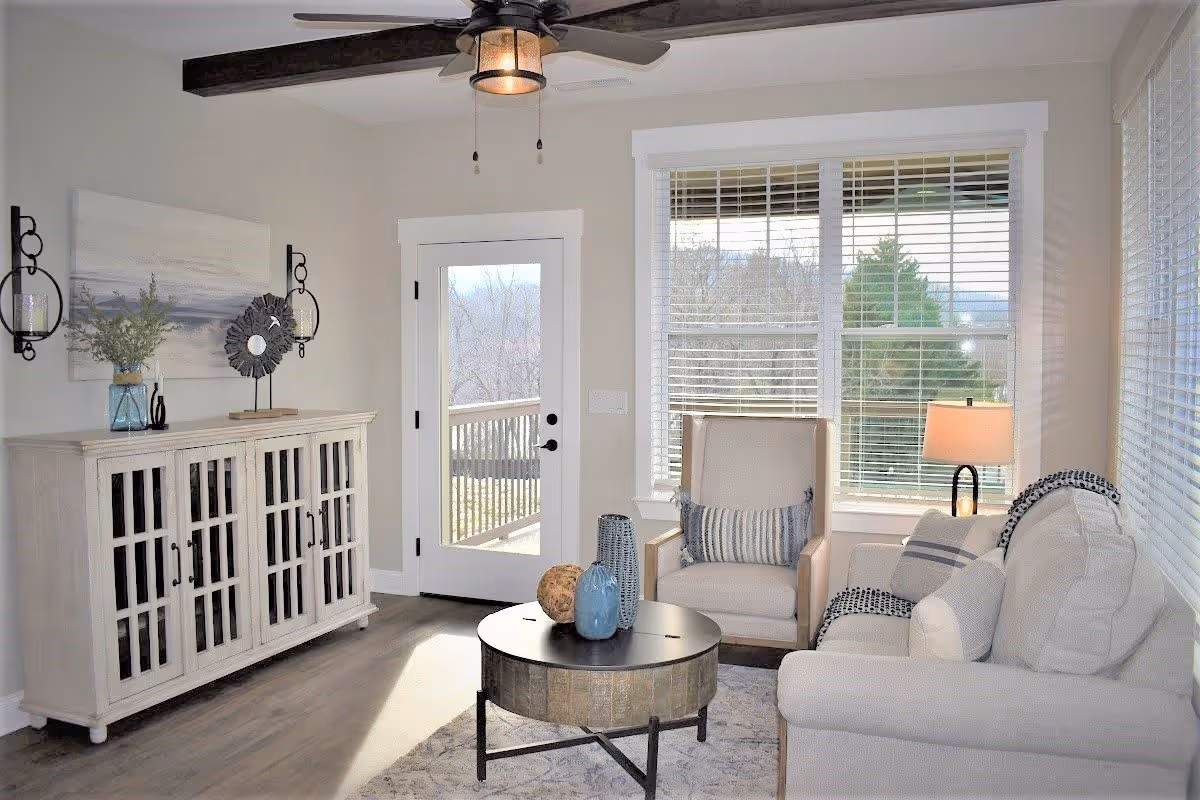 Bright living room with a sofa, armchair, round coffee table, console cabinet, large windows, and a door leading to a deck.