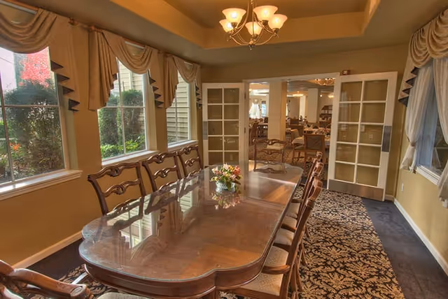 Formal dining room with a long polished wooden table and chairs, chandelier, windows with drapes, and glass-paneled double doors opening to another dining area.