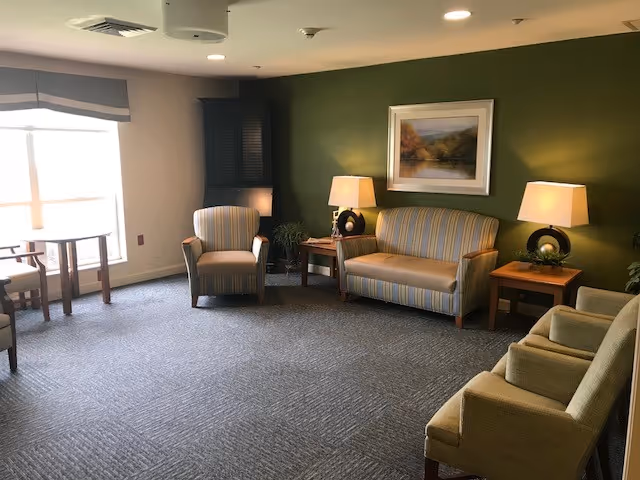 A cozy living room area with a green accent wall featuring a framed landscape painting. The room contains a striped loveseat, two matching armchairs, a wooden side table with a lamp on each side of the loveseat, and a small round table with chairs near a window with a valance. The floor is carpeted in a neutral tone.
