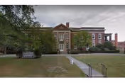 Front exterior view of a large brick building with classical architectural elements, including columns and a pediment. The building is surrounded by trees and a grassy lawn with a paved walkway leading to the entrance.