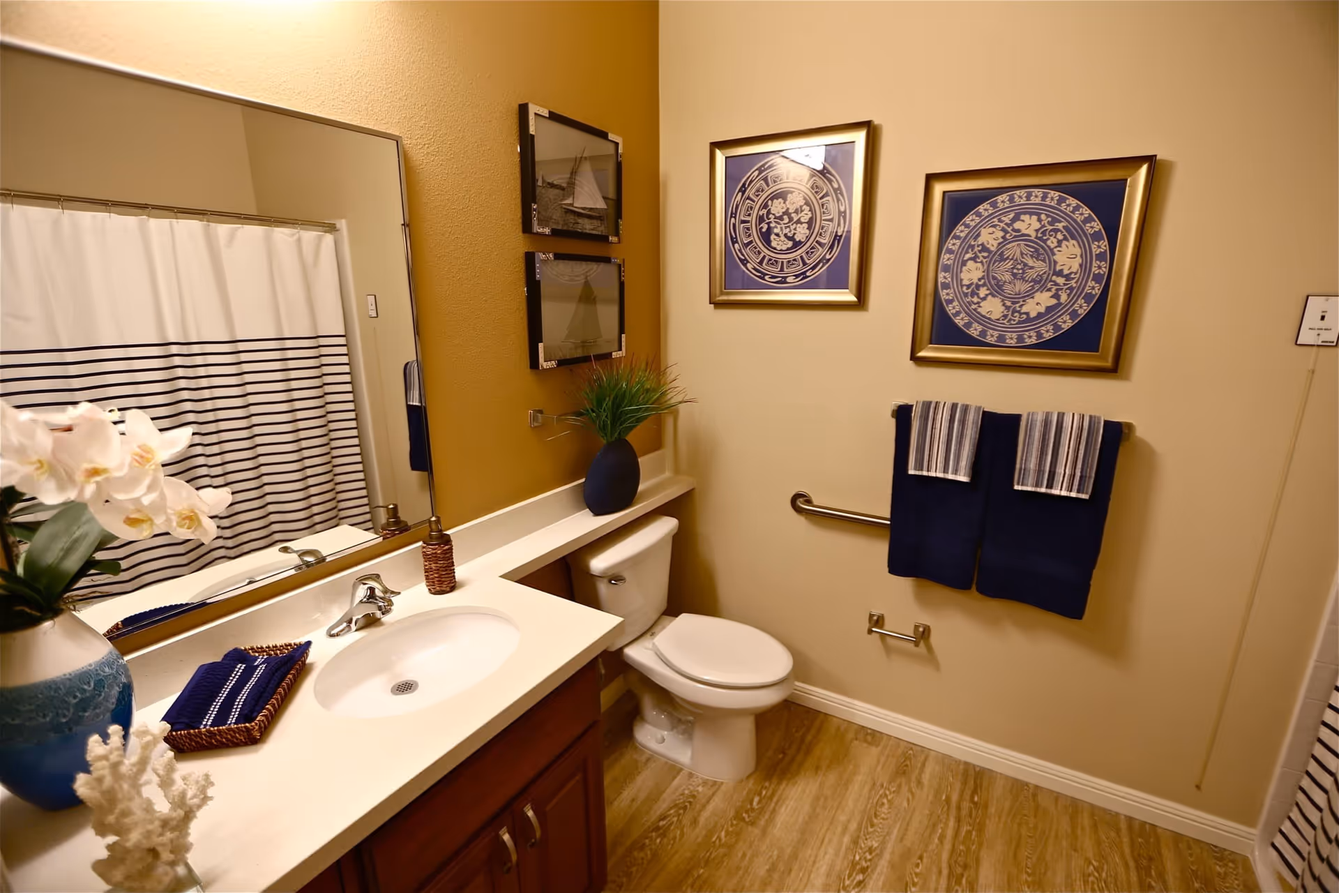 A clean and well-decorated bathroom featuring a white sink with a silver faucet, a large mirror above the sink, a toilet, and a shower with a white curtain that has navy blue stripes. The walls are painted in warm tones with two framed decorative pictures and two navy blue towels hanging on a silver towel rack. There are decorative items including a blue vase with white flowers and a small plant on the countertop.