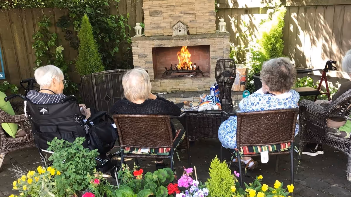 Four elderly individuals sitting outdoors in wicker chairs around a stone fireplace with a fire burning. The area is surrounded by a wooden fence and lush greenery, with colorful flowers in the foreground. Two of the individuals are in wheelchairs, and there are various items on the table in front of the fireplace.