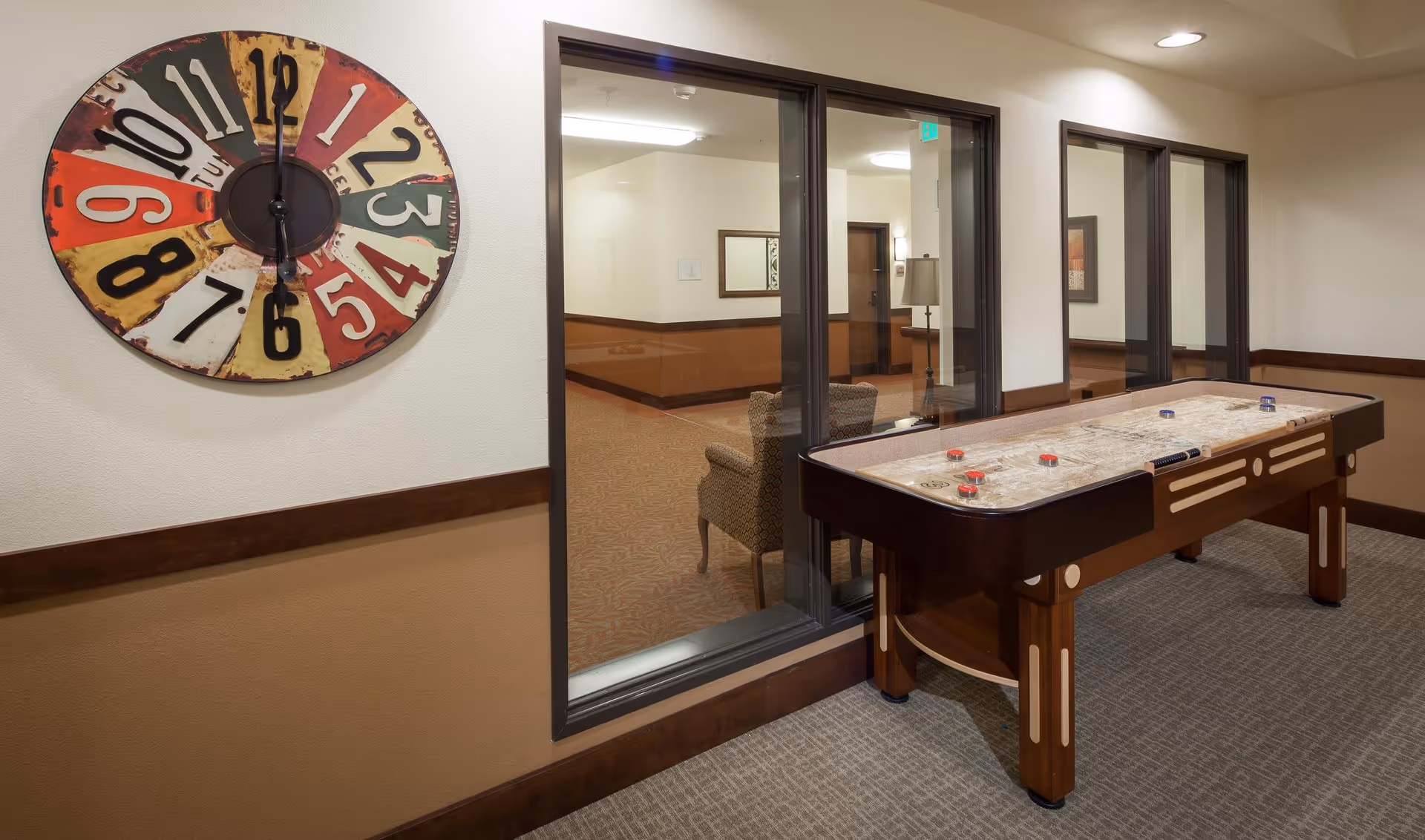 Interior view of a senior living facility game room featuring a shuffleboard table and a large, colorful wall clock. Through the glass window, a seating area with an armchair and a lamp is visible in the adjacent room.
