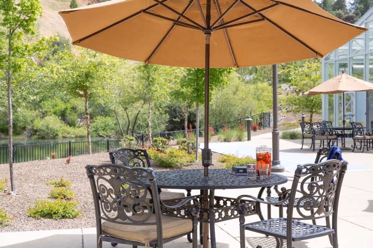 Outdoor patio with ornate metal table and chairs under a large tan umbrella overlooking landscaped grounds and a pond.