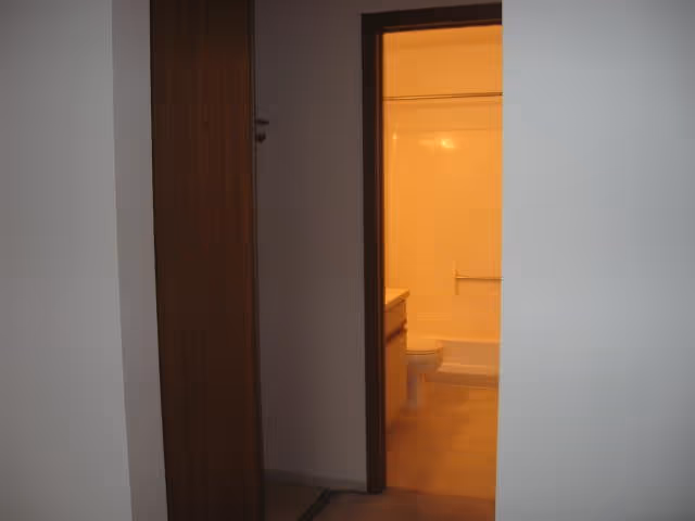 View through a doorway into a bathroom with a toilet, sink, and bathtub with a shower curtain. The bathroom is lit with warm lighting and has beige tile flooring. The surrounding walls outside the bathroom are plain and white.