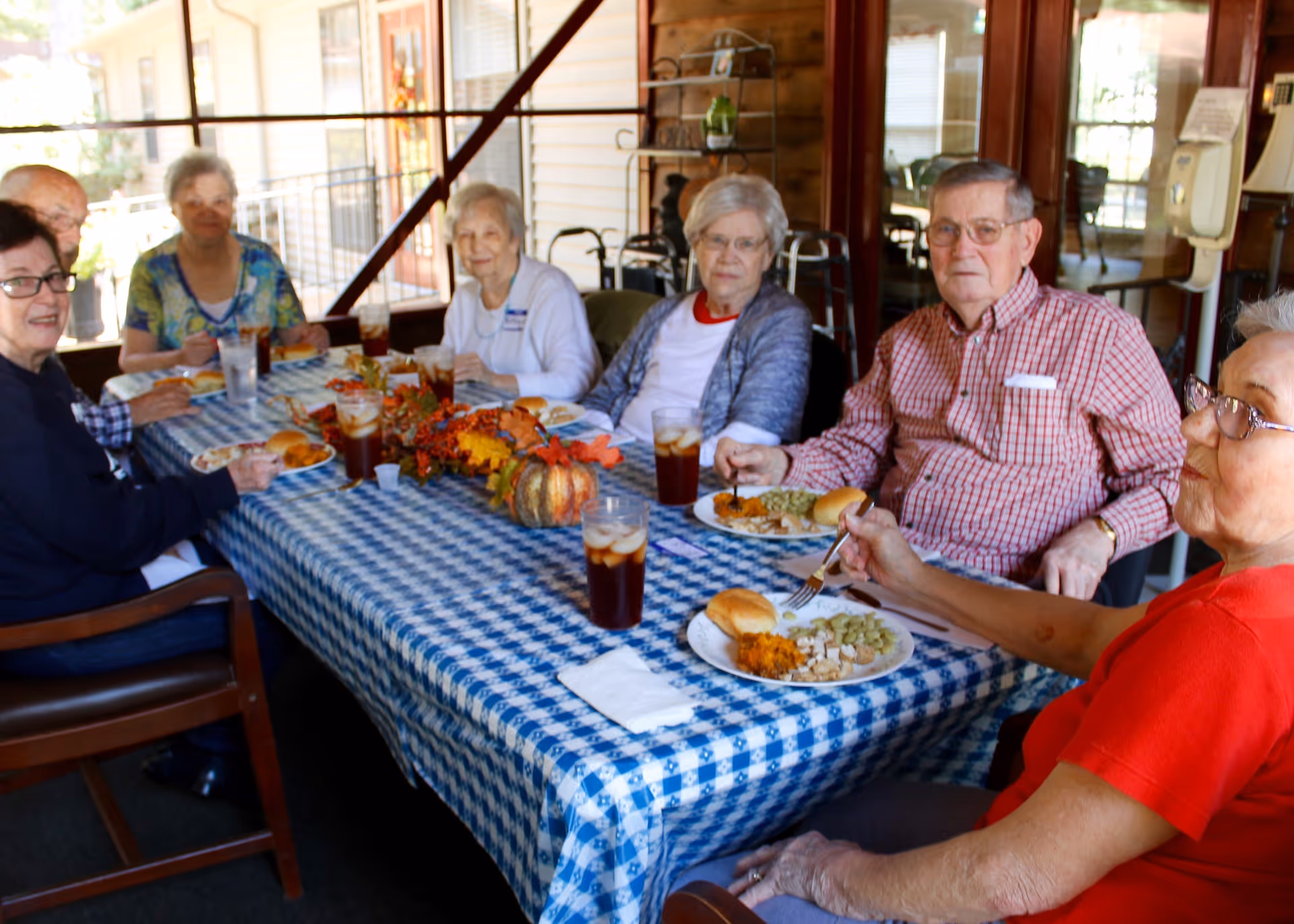 A group of elderly people sitting around a table covered with a blue and white checkered tablecloth, enjoying a meal together. The table is decorated with autumn-themed centerpieces including small pumpkins and leaves. Each person has a plate of food and a glass of iced tea. The setting appears to be a covered porch or patio area with windows and doors in the background.