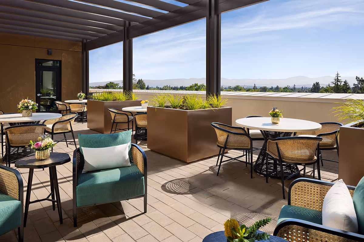 Outdoor patio area with round tables and wicker chairs, green cushioned armchairs with white pillows, large brown planters with green plants, and a pergola overhead. The background shows a clear blue sky and distant hills.