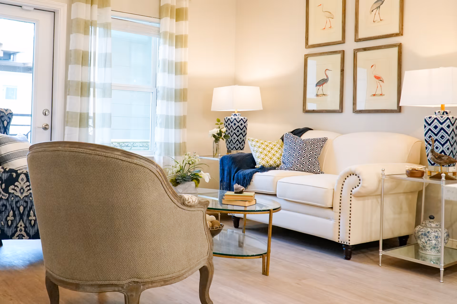 A cozy living room with a white upholstered sofa adorned with patterned pillows and a dark blue throw. There are two matching table lamps with blue and white geometric bases on either side of the sofa. A glass coffee table with books and decorative items sits in front of the sofa. A beige upholstered armchair is positioned in the foreground. The room has light wood flooring, a window with striped curtains, and four framed bird illustrations on the wall.