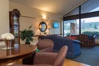 A cozy living room area with a round wooden table and two brown chairs in the foreground, a blue couch and a striped armchair near large windows letting in natural light. A grandfather clock, a green plant, a wall-mounted TV, and a round mirror with a lamp underneath are visible on the wall.