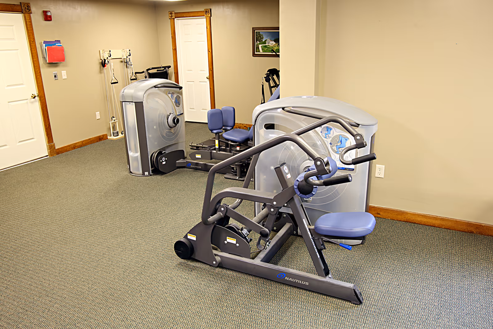 A small exercise room with two Nautilus strength training machines on a carpeted floor. The walls are beige with wooden trim, and there are two white doors and a framed picture on the wall.