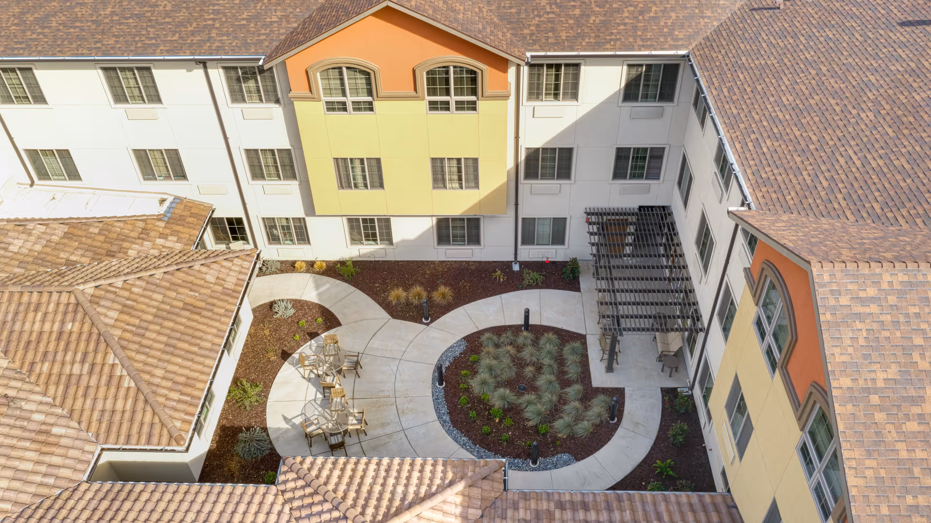 Aerial view of a senior living facility courtyard with a circular concrete pathway, outdoor seating with tables and chairs, landscaped garden beds, and a pergola attached to the building. The building has multiple windows and a tiled roof with sections painted in white, yellow, and orange.