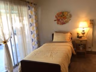 A small bedroom with a single bed covered in beige bedding. There is a nightstand with a lamp and some decorative items next to the bed. A colorful wall decoration hangs above the bed. To the left, there are large glass sliding doors with patterned curtains allowing natural light into the room.